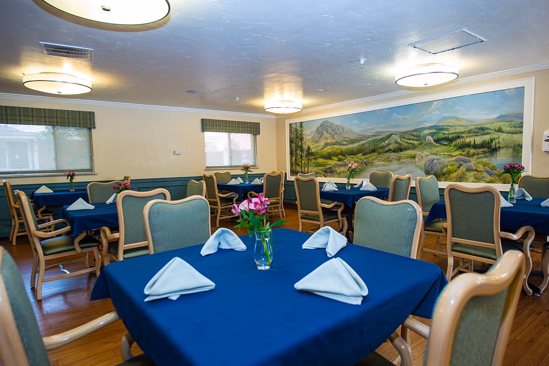 A dining room with several tables covered in blue tablecloths, each set with folded white napkins and a small vase of pink flowers. The room has green cushioned chairs with wooden frames, two windows with green valances, and a large mural of a scenic mountain landscape on one wall. The ceiling has round light fixtures providing warm lighting.