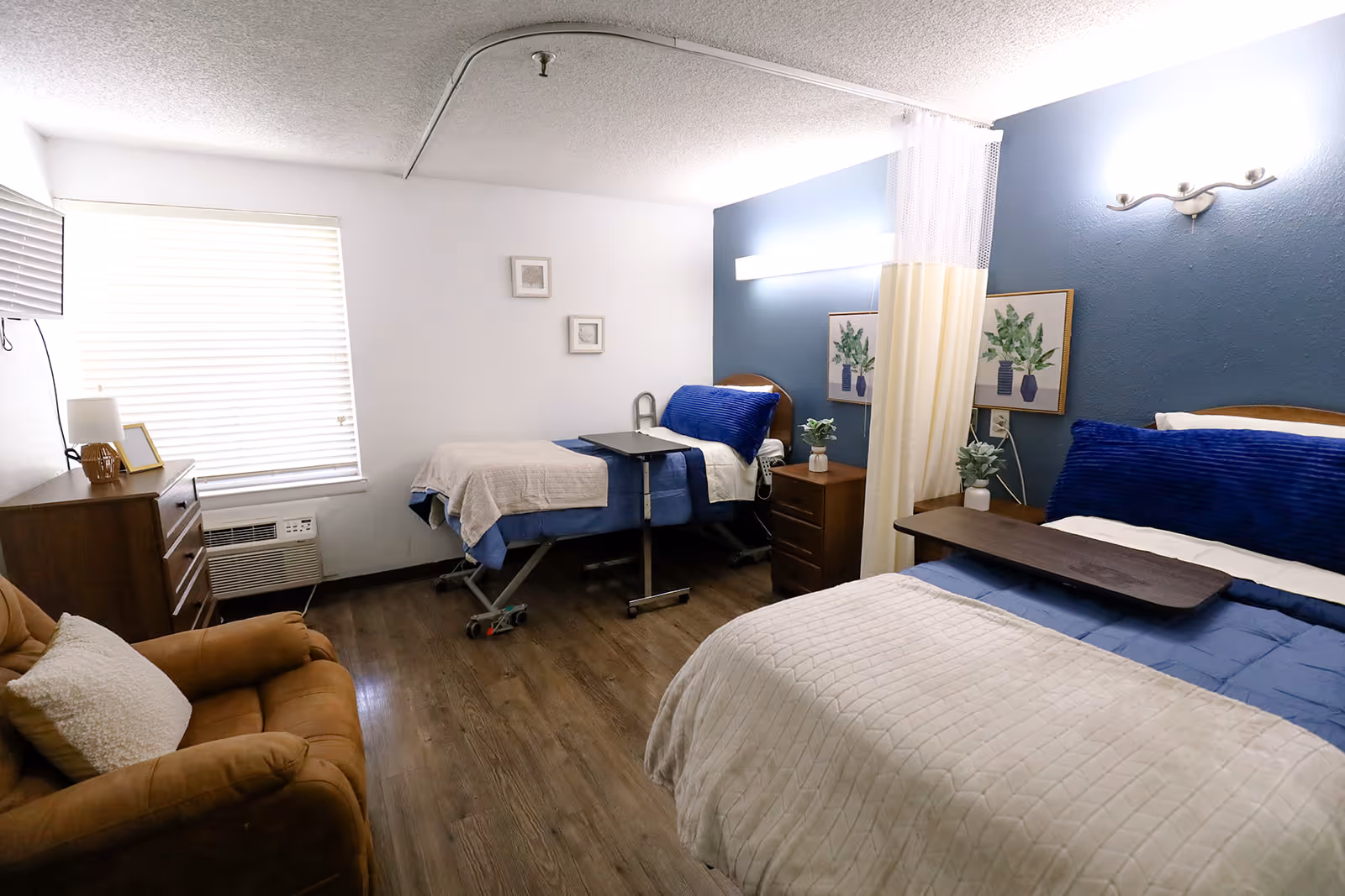 A shared bedroom in a healthcare facility with two beds, one with a blue headboard and the other with a beige blanket. Each bed has a small overbed table. There is a brown armchair with a white pillow, a wooden dresser with a lamp and picture frame, and a window with blinds. The walls are white and blue, decorated with framed botanical prints and small plants on nightstands. A privacy curtain is partially drawn between the beds.