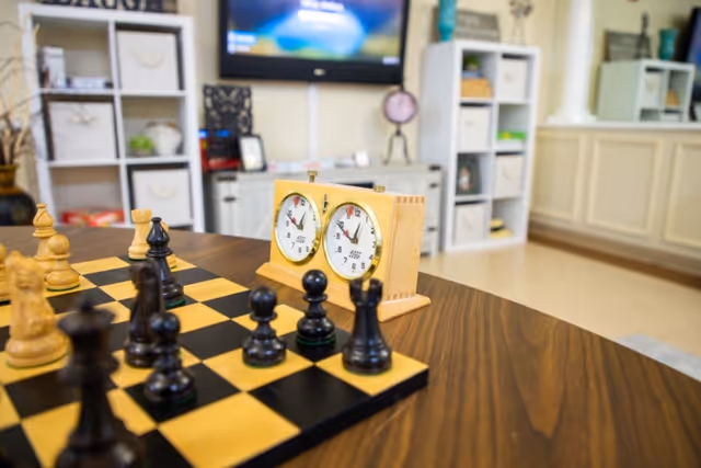 A close-up view of a chessboard with black and white chess pieces set up for a game on a wooden table. Behind the chessboard is a wooden chess clock. In the background, there are shelves with various items and a television mounted on the wall in a well-lit room.