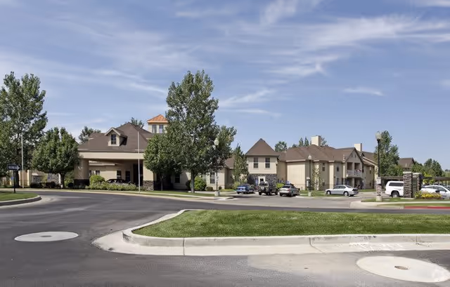 Exterior view of a senior living facility named Rose Cove Apartments with a paved driveway, parked cars, green trees, and a clear blue sky.