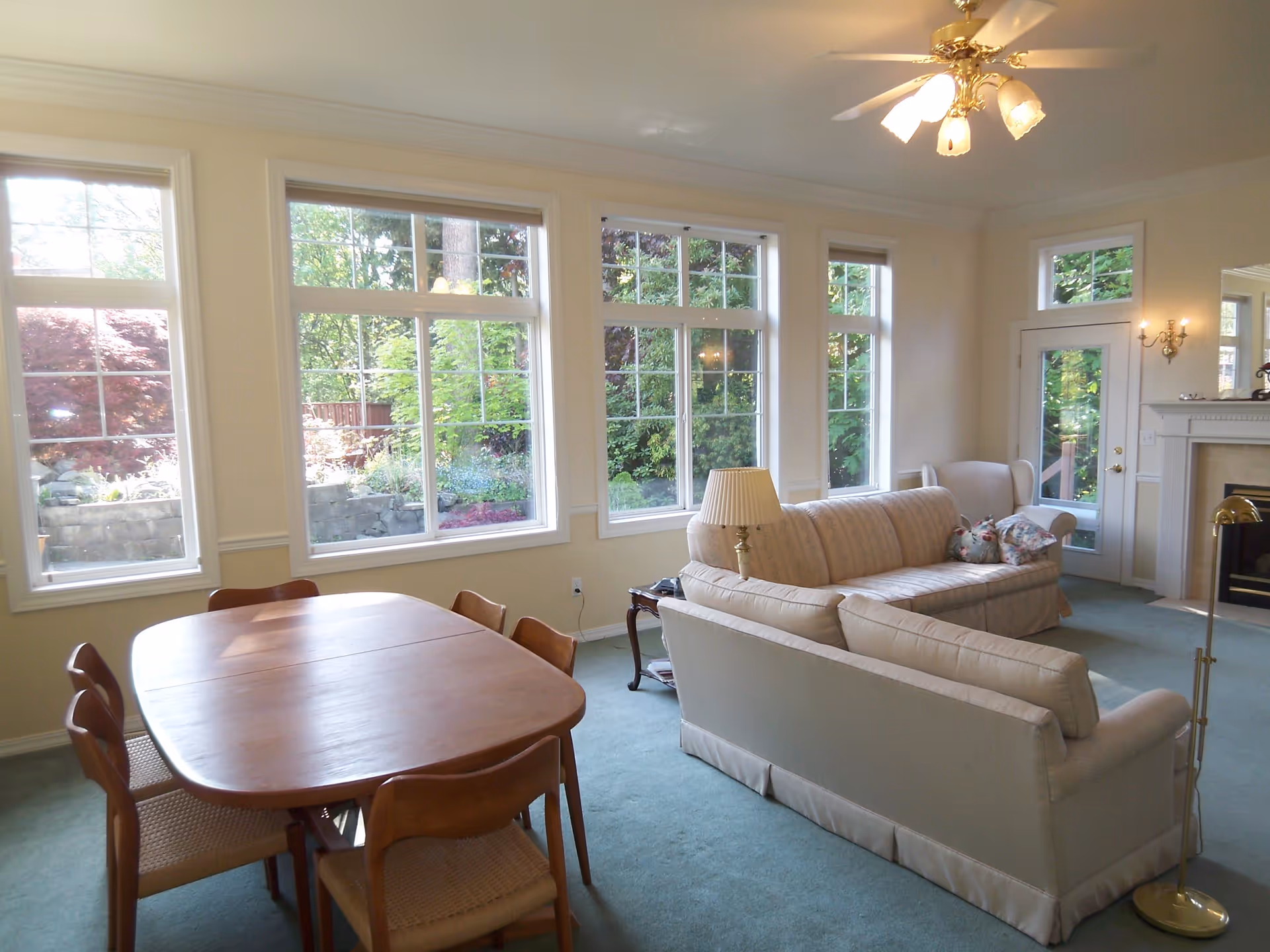 A bright living room with large windows showing a garden outside. The room has a wooden dining table with six chairs on the left, two beige sofas with floral pillows in the center, a floor lamp, a table lamp, a fireplace on the right, and a ceiling fan with lights.