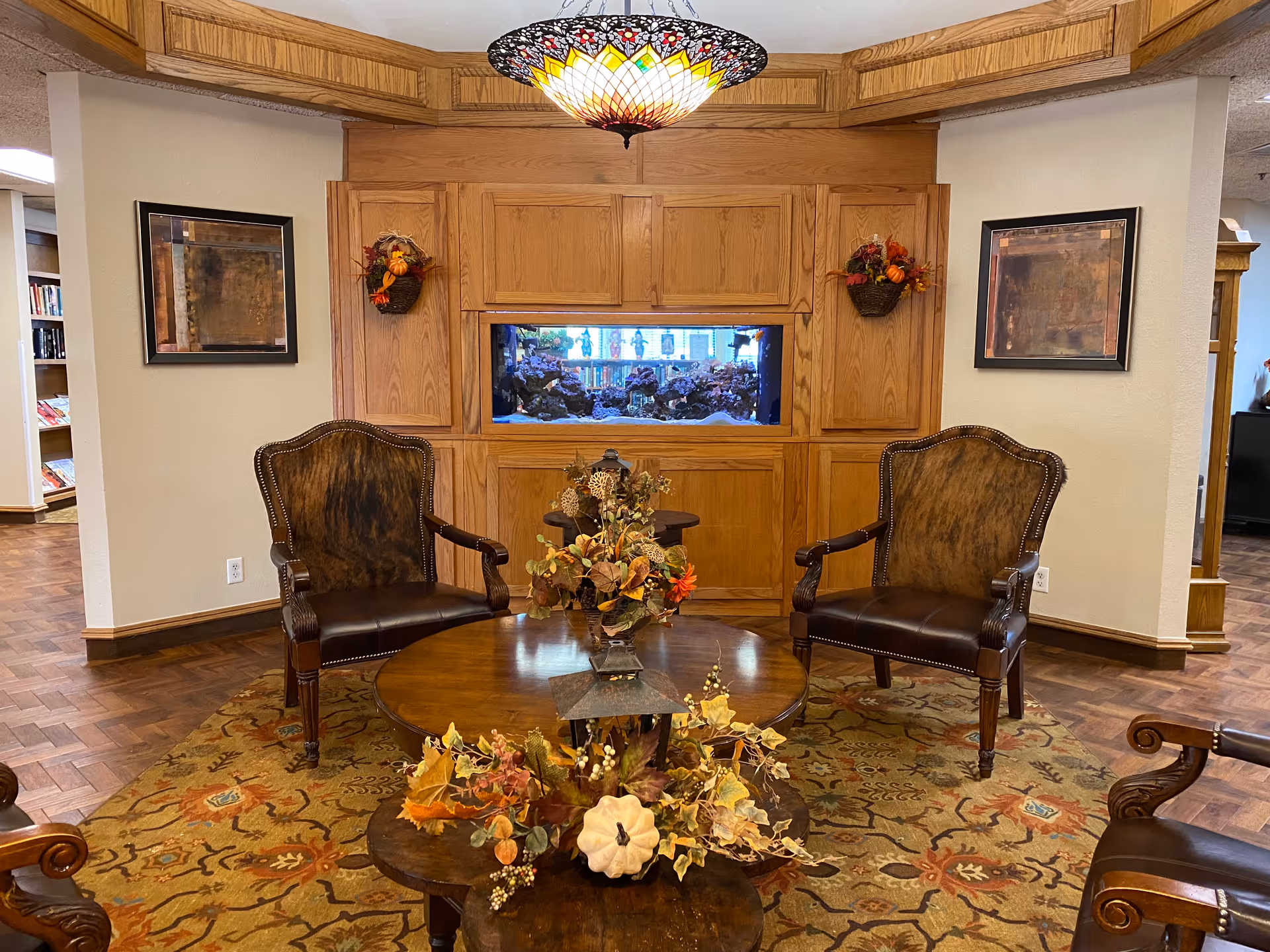 A cozy seating area in a senior living facility with two brown leather and wood armchairs facing a round wooden coffee table decorated with autumn-themed floral arrangements. Behind the chairs is a built-in wooden cabinet with an aquarium in the center, flanked by two framed abstract paintings and small wall-mounted baskets with fall decorations. The floor has a patterned area rug, and a colorful stained glass ceiling light fixture hangs above.