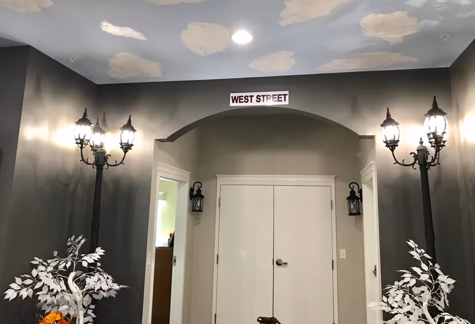 Interior hallway with double doors under an arch labeled "West Street", flanked by decorative street lamps and white potted plants.