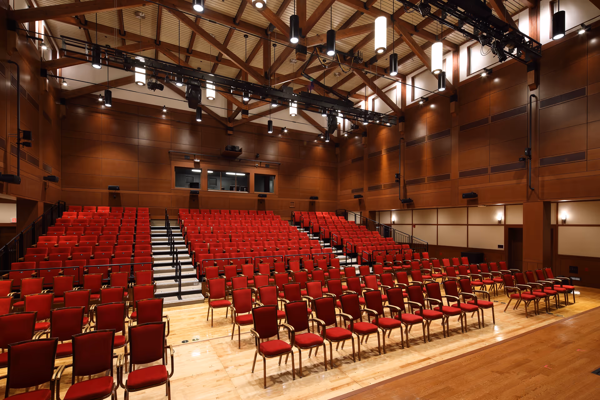 Empty auditorium with rows of red seats facing a wooden stage under exposed beam ceiling and stage lighting.