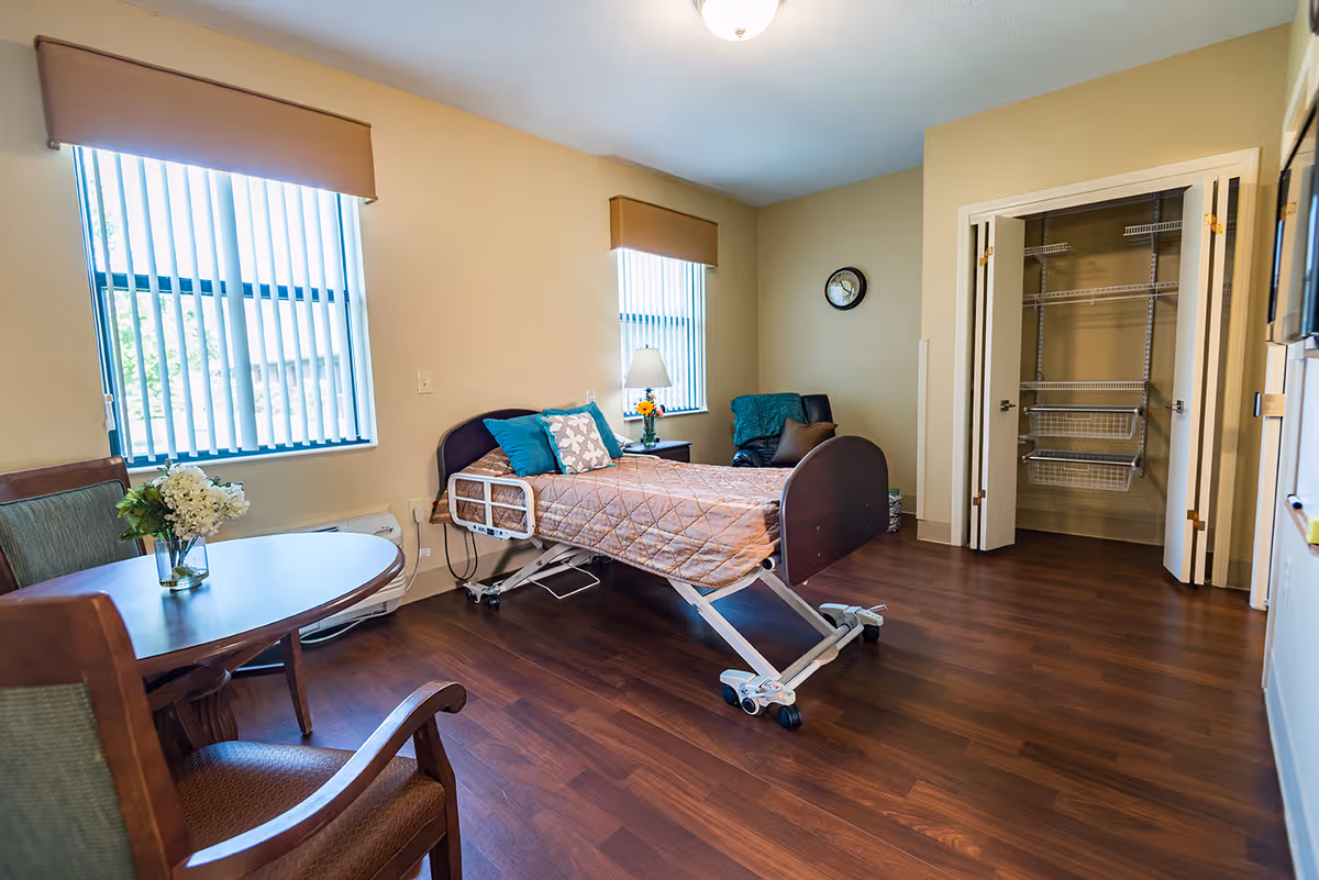 A well-lit room with a hospital-style bed covered with a quilt and pillows, two windows with vertical blinds, a round wooden table with two chairs, a small side table with a lamp and flowers, an armchair with a blanket, and an open closet with wire shelving. The floor is dark wood and the walls are painted beige.