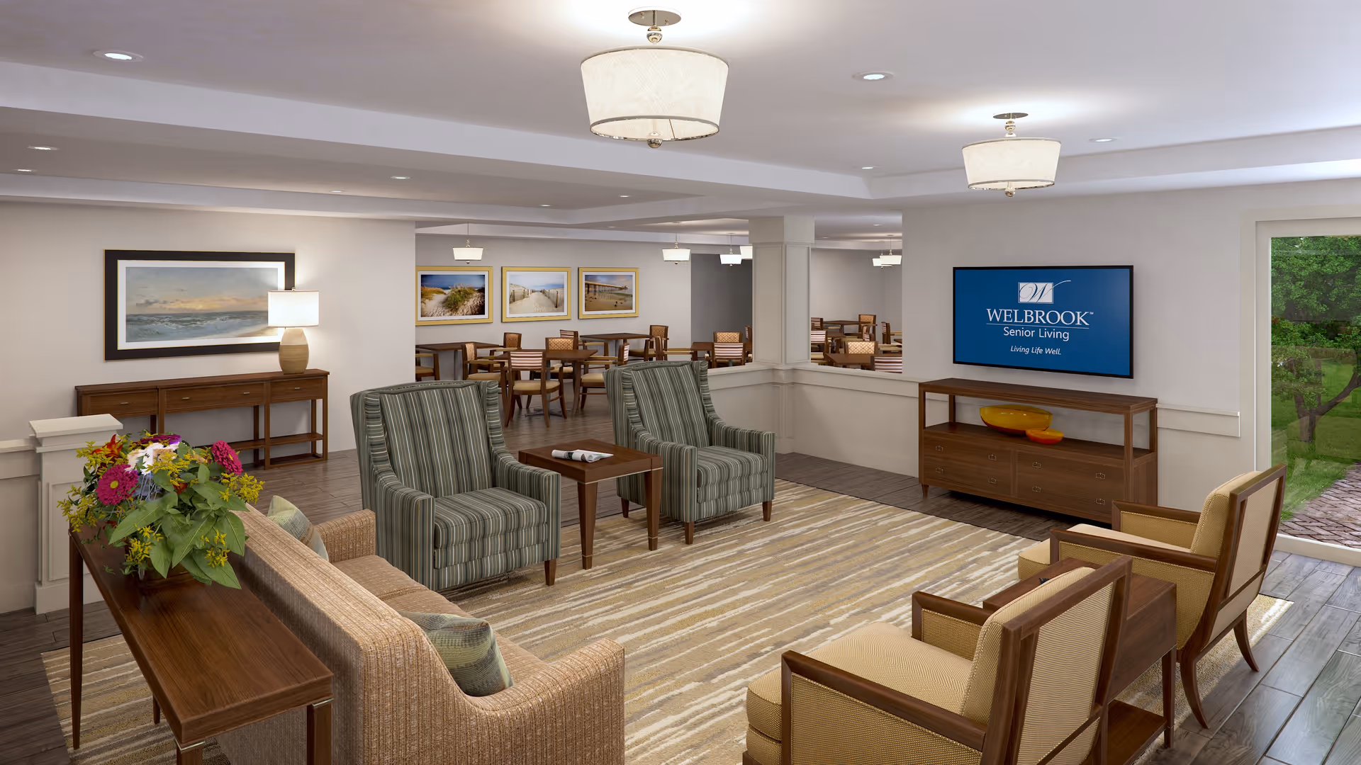 A well-lit senior living facility common area with comfortable seating including a sofa, striped armchairs, and beige chairs arranged around a wooden coffee table on a patterned rug. The room features wooden furniture, framed landscape artwork on the walls, a flat-screen TV displaying the Welbrook Senior Living logo, and a large window showing a green outdoor space.