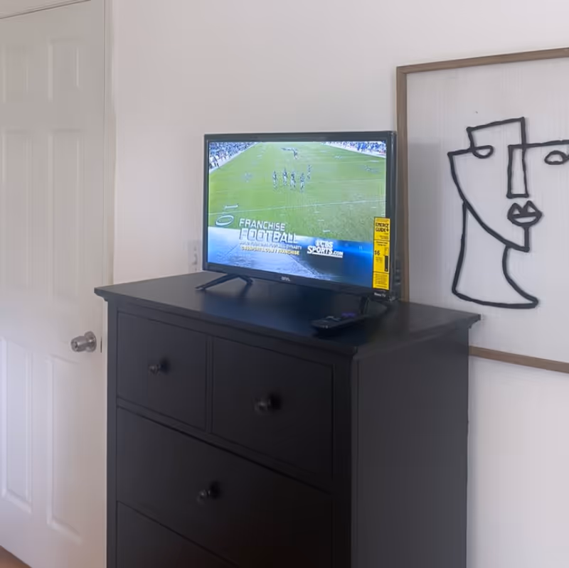 A black dresser with four drawers stands against a white wall. On top of the dresser is a flat-screen TV displaying a football game. A remote control is placed next to the TV. To the right of the dresser, a framed abstract line art of a face hangs on the wall. A white door with a silver doorknob is partially visible on the left side of the image.