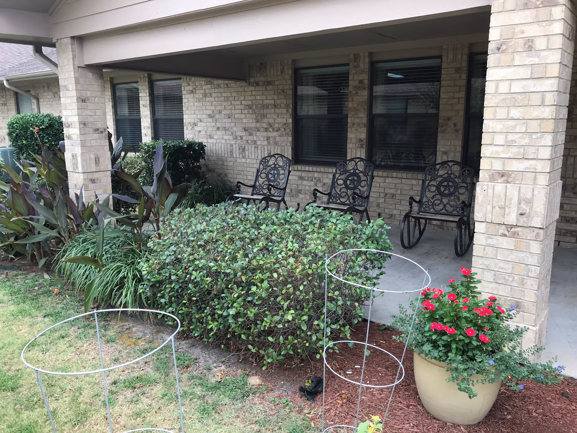 Covered brick porch with three decorative metal rocking chairs, shrubs, and a potted flowering plant in front of the building.