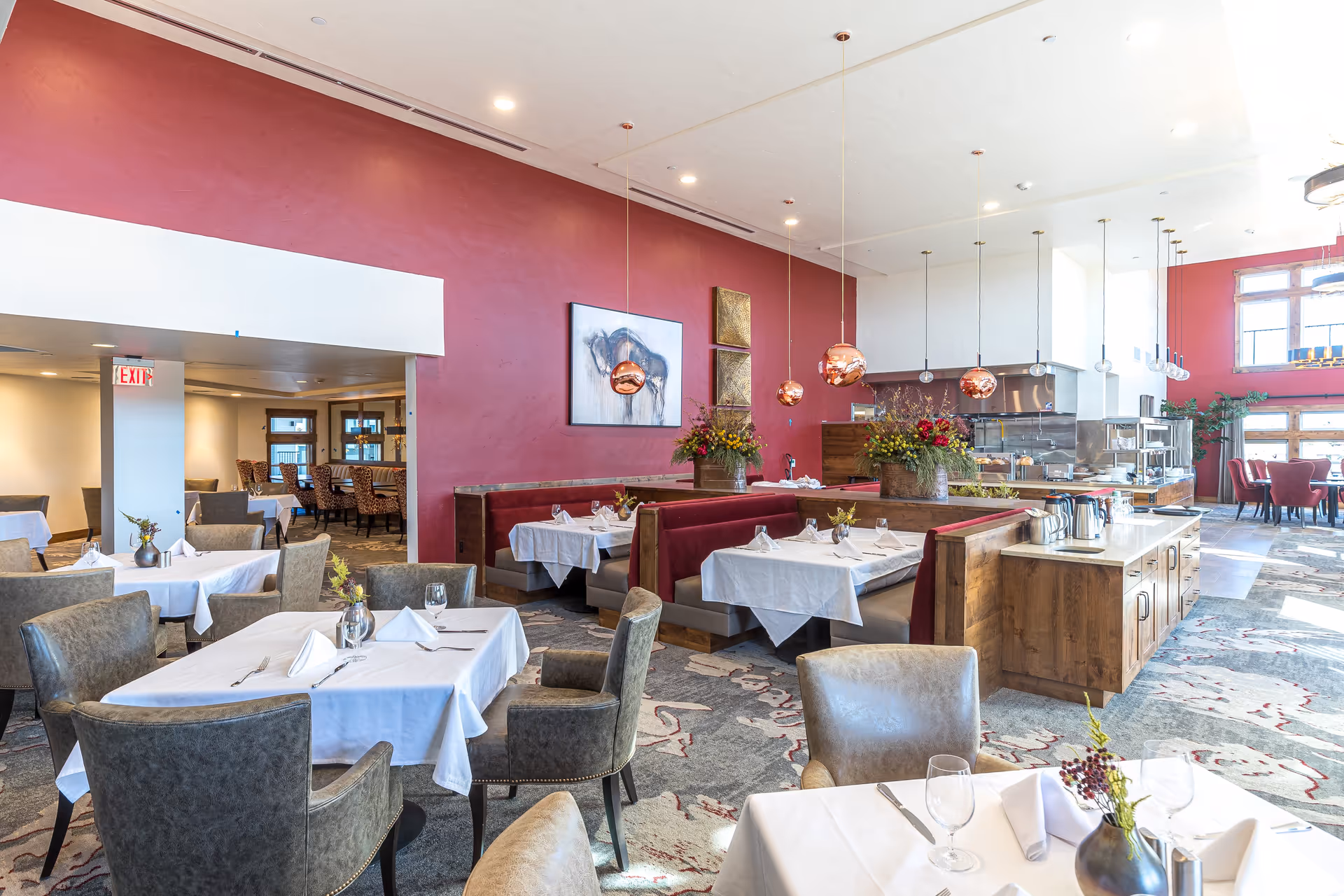 A bright and spacious dining room with tables covered in white tablecloths, set with napkins, glasses, and silverware. The room features comfortable upholstered chairs and red booth seating along a red accent wall. Copper pendant lights hang from the ceiling, and there are floral arrangements on the tables and counters. Large windows allow natural light to fill the space.