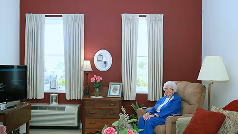 A senior woman with white hair and glasses sitting in a beige recliner chair in a cozy living room. The room has a red accent wall with two windows covered by beige curtains. Between the windows is a wooden chest of drawers with a lamp, a vase of pink flowers, and framed pictures. A television is on a stand to the left, and a table with a floral arrangement is in the foreground.