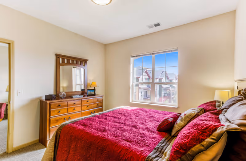Sunlit bedroom with a red quilted bed, wooden dresser and mirror, and a window showing neighboring townhouses.