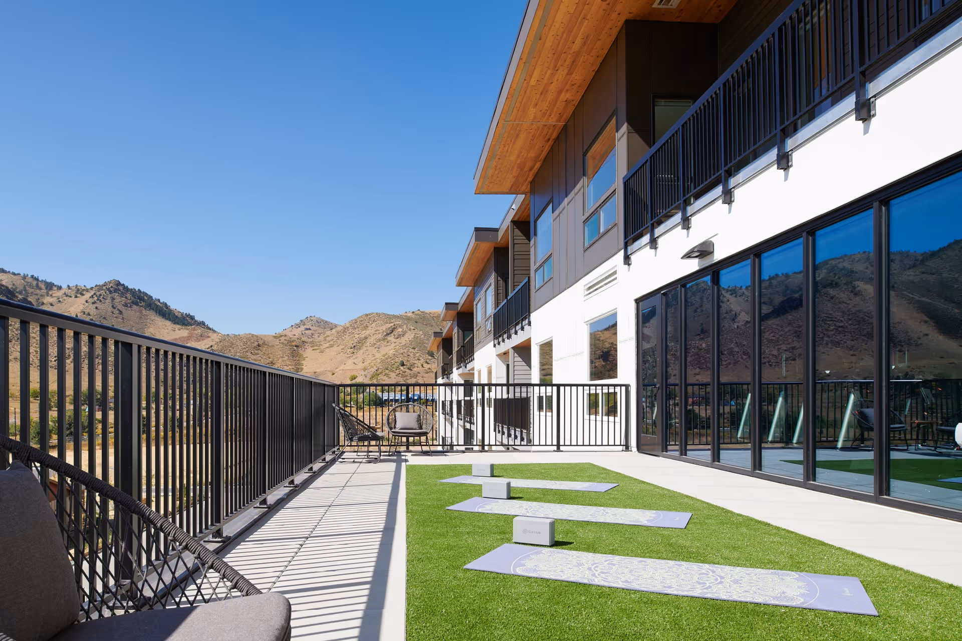 Outdoor patio area at a senior living facility with artificial grass and yoga mats laid out for exercise. The patio has black metal railings and modern outdoor seating. The building has large glass windows reflecting the surrounding mountainous landscape under a clear blue sky.