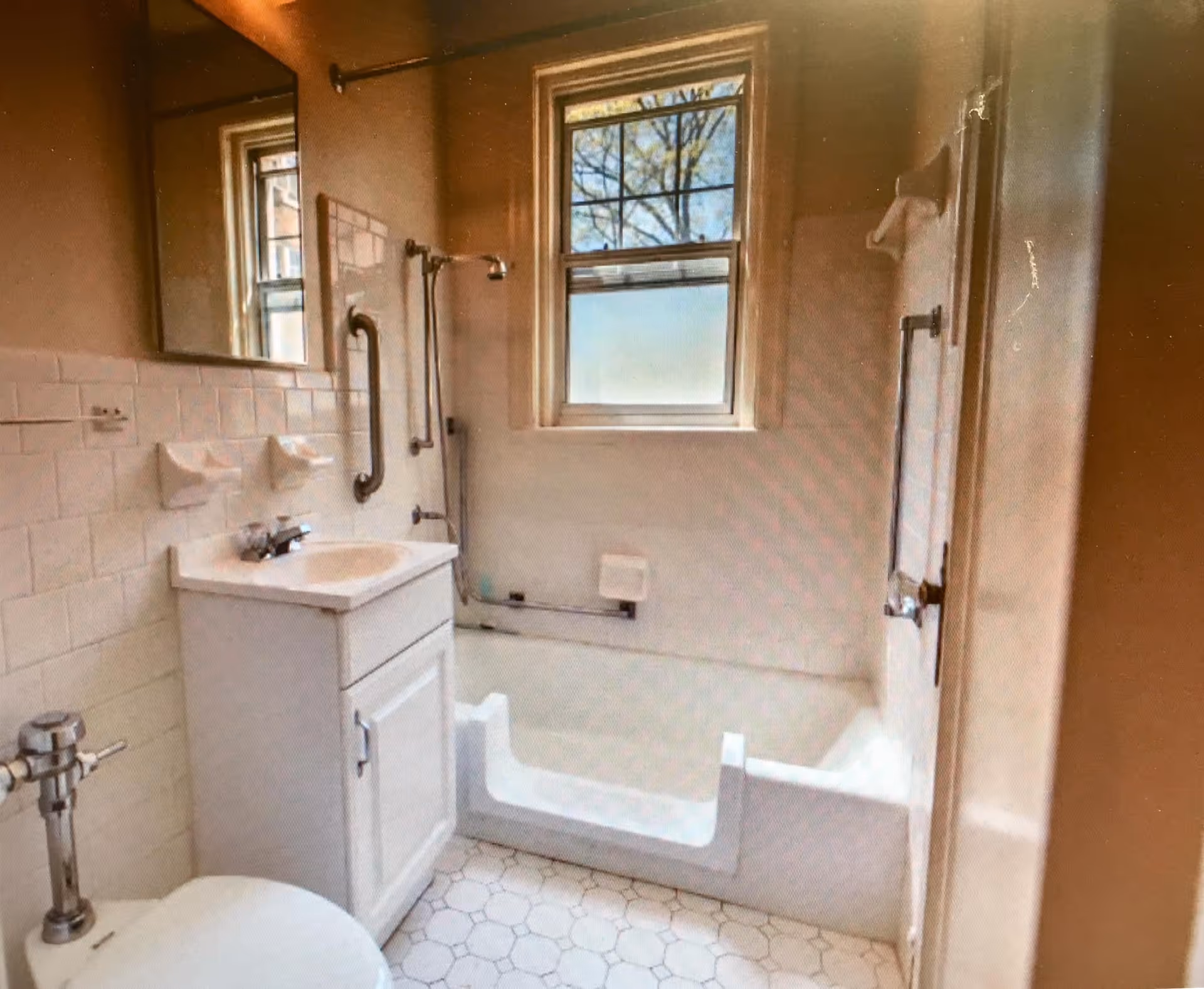 A small bathroom with a white toilet, a white vanity with a sink, a large mirror above the sink, and a bathtub with a cut-out entry for accessibility. There is a window above the bathtub letting in natural light, and grab bars are installed near the bathtub and shower area.