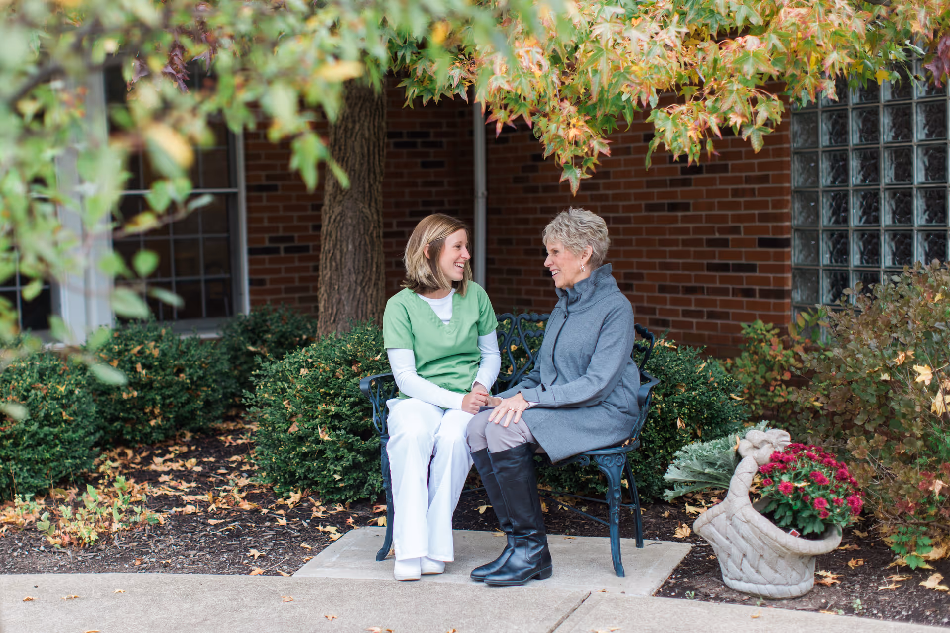 Two women sitting on a bench outside near bushes and a brick building. One woman is wearing a green scrub top and white pants, and the other woman is wearing a gray coat and black boots. They are smiling and holding hands, engaged in conversation. There is a decorative basket with flowers next to them.