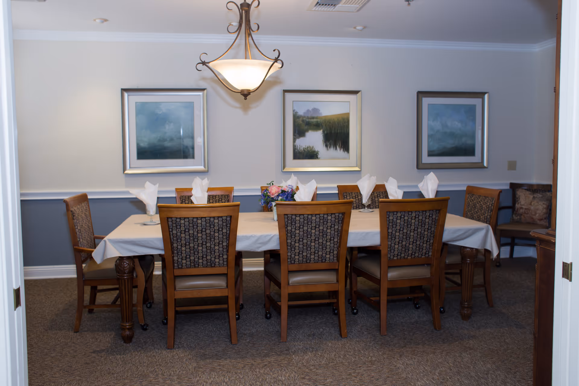 A dining room with a rectangular table covered with a white tablecloth, surrounded by eight wooden chairs with patterned backs. Each place setting has a folded white napkin in a glass. A floral centerpiece is on the table. The walls are painted light gray with a white chair rail, and three framed landscape paintings hang on the wall. A decorative ceiling light fixture hangs above the table.