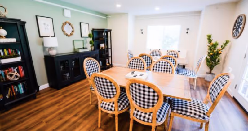 A bright dining room with a wooden table surrounded by eight chairs upholstered in black and white checkered fabric. The room has wooden flooring, a large window letting in natural light, two black bookshelves, a black sideboard with decorative items, framed artwork on the walls, a large clock, and a potted plant near the door.