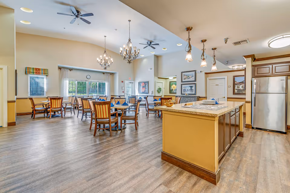 A spacious dining room with multiple wooden tables and chairs arranged neatly. The room features a large kitchen island with a granite countertop, pendant lights hanging above, and a stainless steel refrigerator. The walls are decorated with framed artwork, and large windows allow natural light to fill the space. The ceiling has chandeliers and ceiling fans.