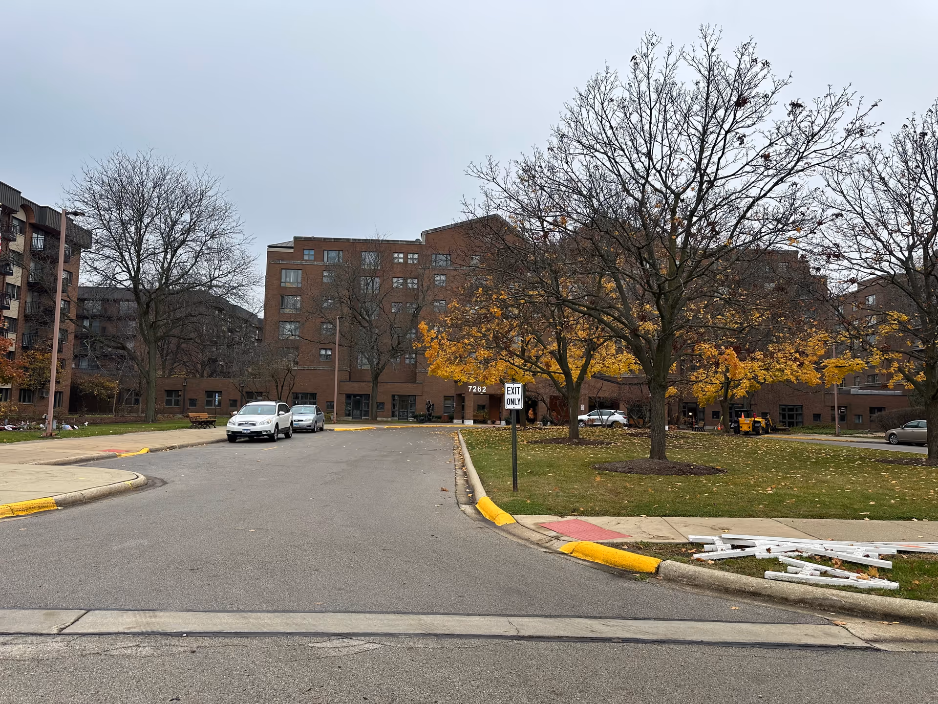 Exterior view of a multi-story brick building with several windows, surrounded by leafless trees and some with yellow autumn leaves. There are a few parked cars along the driveway leading to the building entrance. A sign reads 'EXIT ONLY'. The sky is overcast.