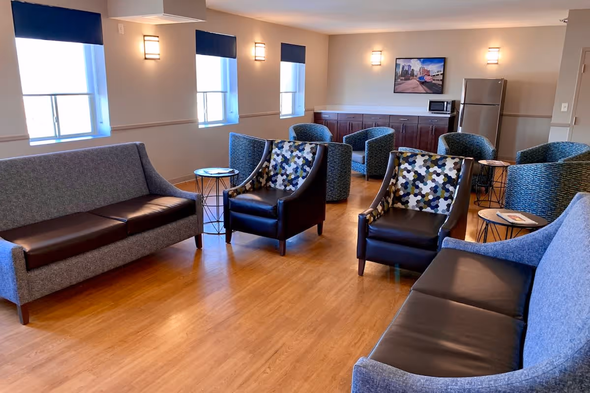 A spacious lounge area with wooden flooring featuring two gray sofas with black cushions, two patterned armchairs, and several blue armchairs arranged around small round tables. The room has three windows with blue shades, wall-mounted lights, a refrigerator, microwave, and a framed picture on the wall.