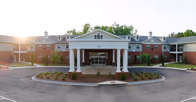 Front exterior view of a two-story senior living facility building with a covered entrance supported by white columns, surrounded by landscaped greenery and a circular driveway.