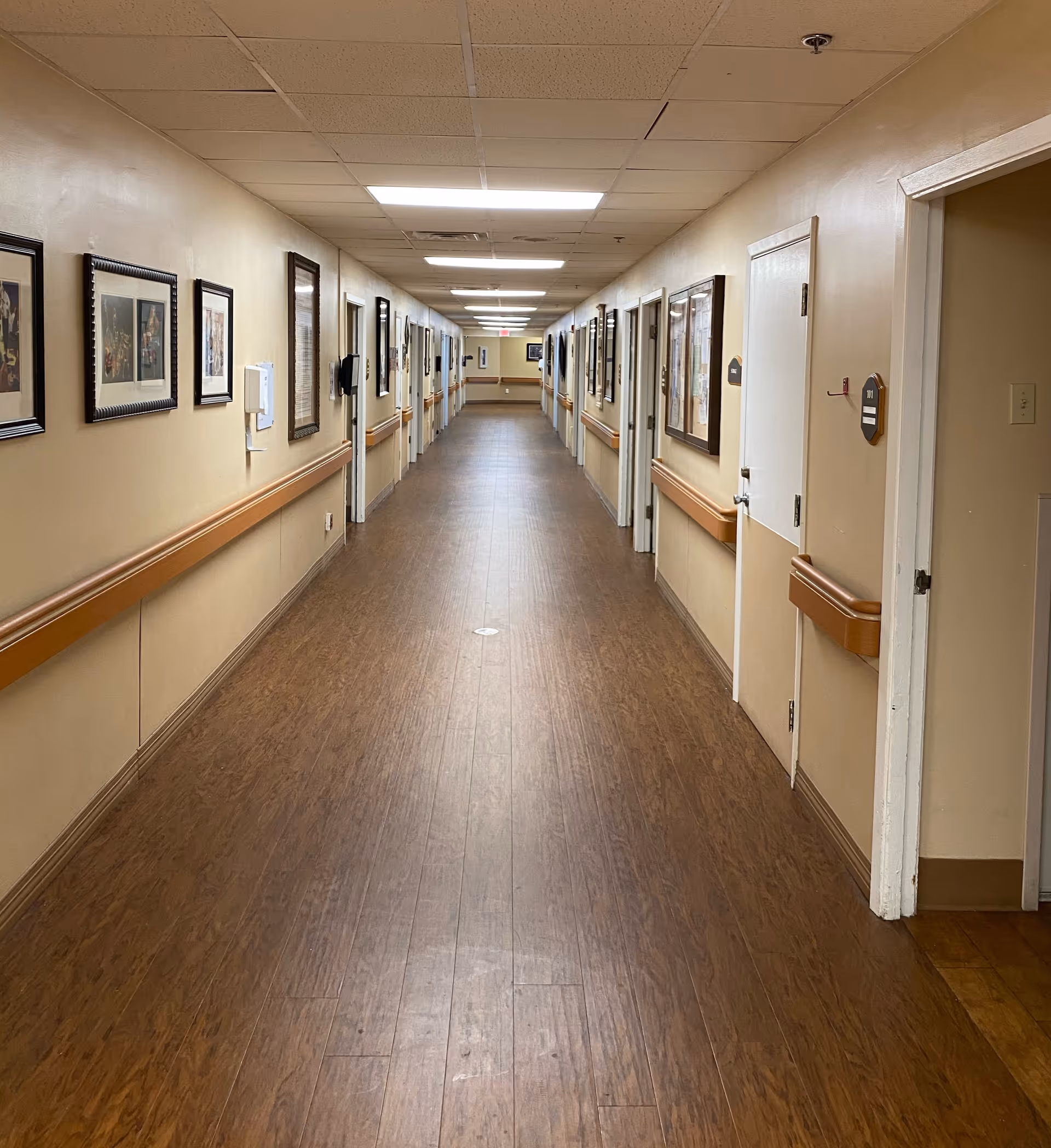 A long, well-lit hallway in a healthcare facility with wood-patterned flooring and beige walls. The hallway is lined with multiple closed doors on both sides, each with handrails beneath the framed pictures and bulletin boards hanging on the walls. The ceiling has recessed lighting panels.