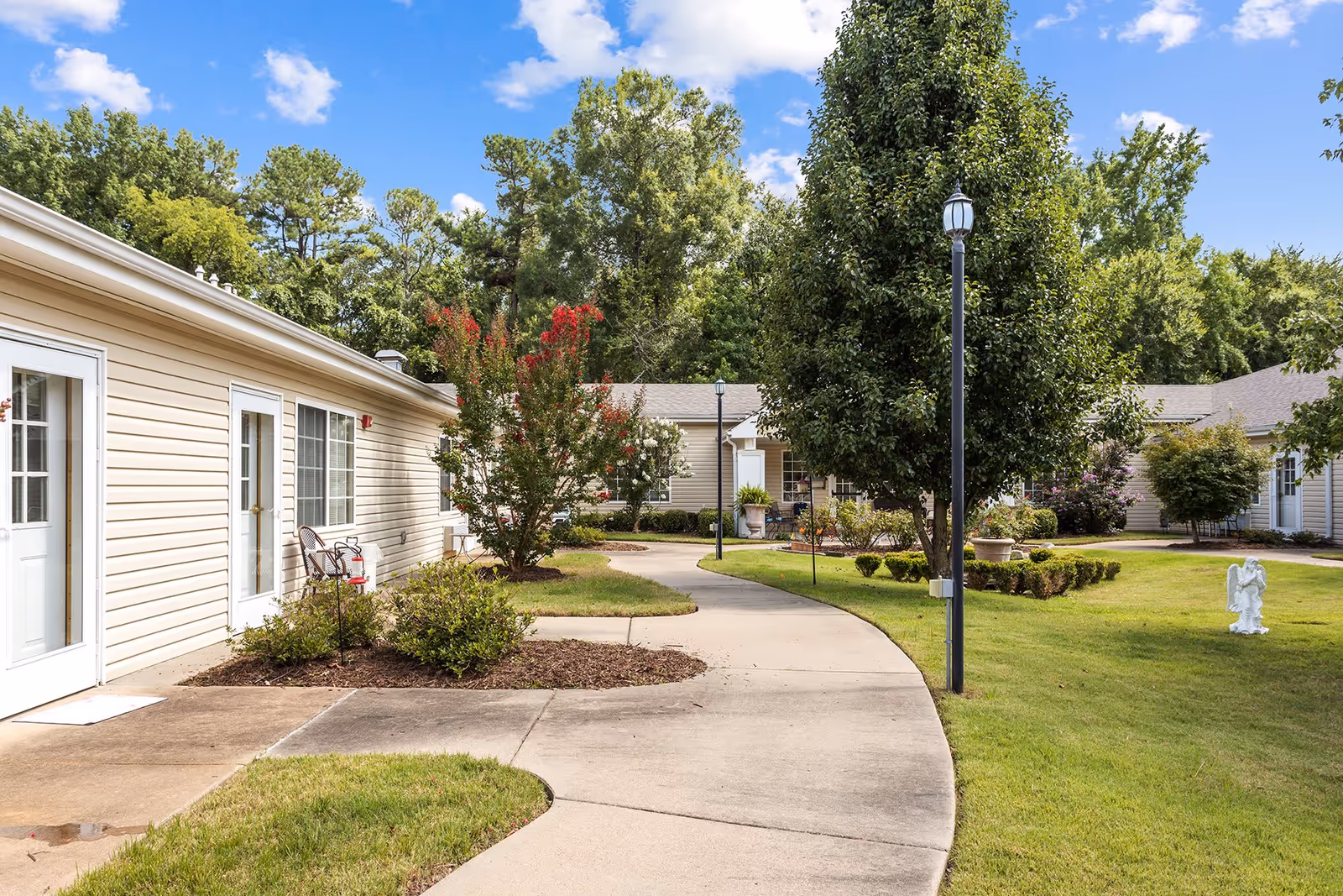 Curved paved walkway through a landscaped courtyard with lamp posts, trees, and single-story beige assisted living buildings.