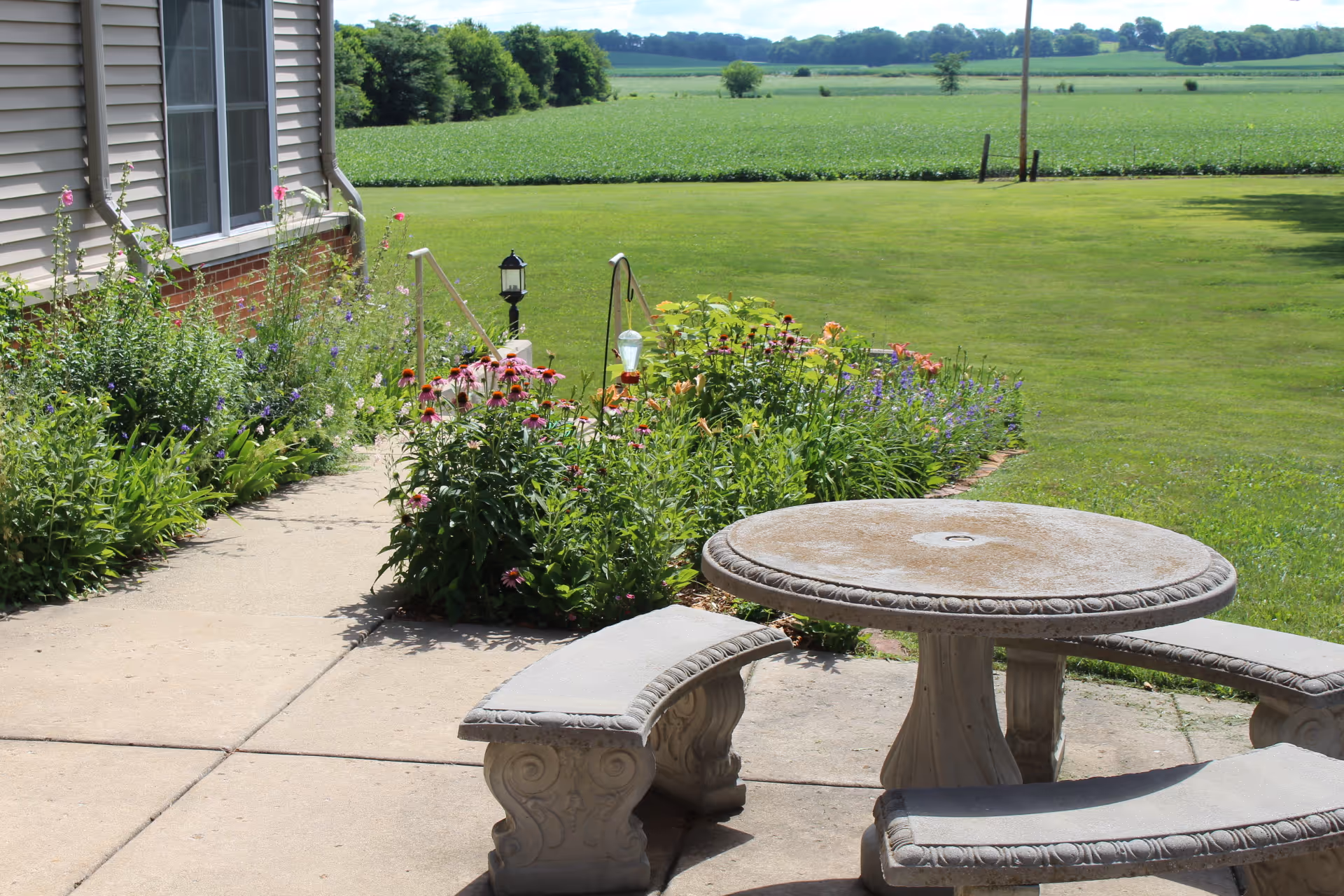 Outdoor patio area with a round stone table and curved stone benches on a concrete surface. There are flower beds with various blooming flowers along the side of a building, and a large green lawn and fields extend into the distance under a clear sky.