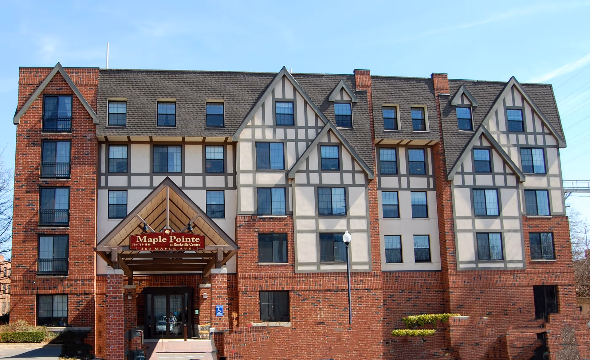 Front exterior view of Maple Pointe Senior Living building, a multi-story structure with a combination of brick and Tudor-style facade, multiple windows, and a covered entrance with a sign displaying the facility name.