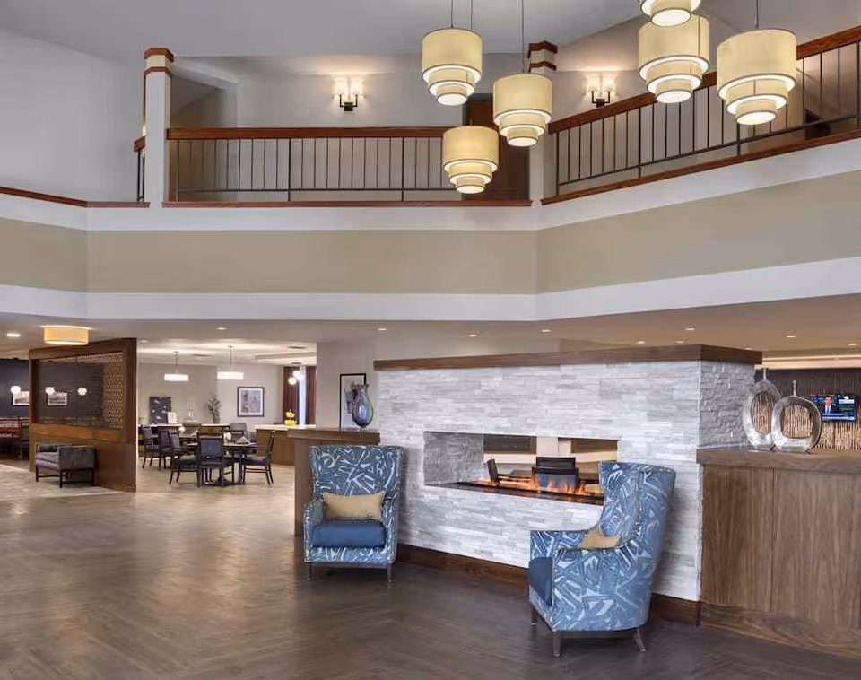 Spacious and modern interior of a senior living facility featuring a double-sided stone fireplace with two blue patterned armchairs in front. The area has high ceilings with multiple hanging pendant lights and a balcony railing above. In the background, there are dining tables and chairs, and a reception or lounge area with decorative items and a television.