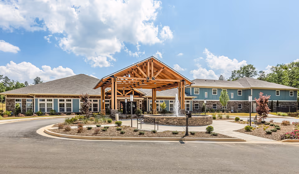 Front exterior view of Oaks at Eagle's Landing facility featuring a large covered entrance with wooden beams, a circular driveway, landscaped garden beds, and a water fountain under a partly cloudy sky.