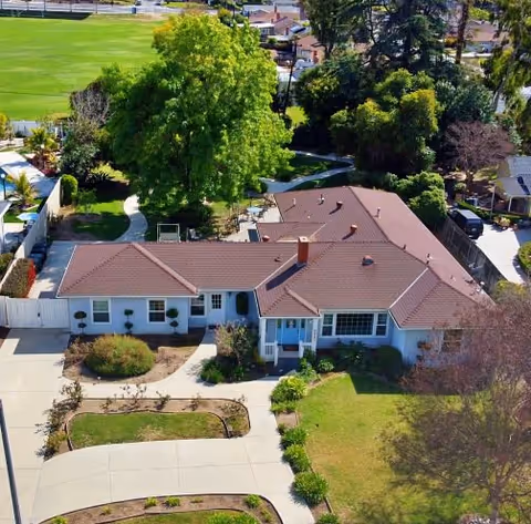 Aerial view of a single-story residential building with a tiled roof, driveway, lawns and surrounding trees.