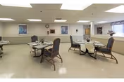 A dining room with several tables and chairs arranged for meals. The room has a tiled floor, white walls with a chair rail, and fluorescent ceiling lights. There are framed pictures on the walls and a clock above a doorway. A large window with a valance lets in natural light.