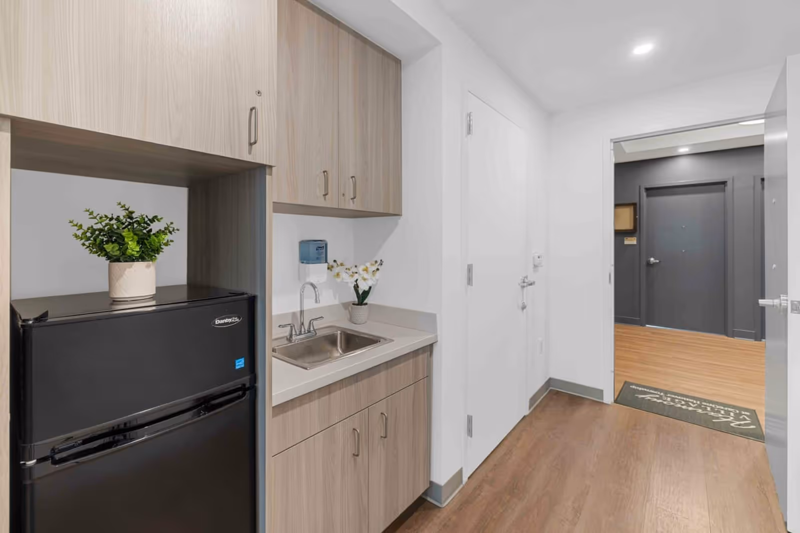 Small kitchenette area with light wood cabinets, a black mini refrigerator with a potted plant on top, a stainless steel sink, and a small potted flower on the countertop. The kitchenette is adjacent to a hallway with wooden flooring and white walls, leading to a dark gray door in the background.