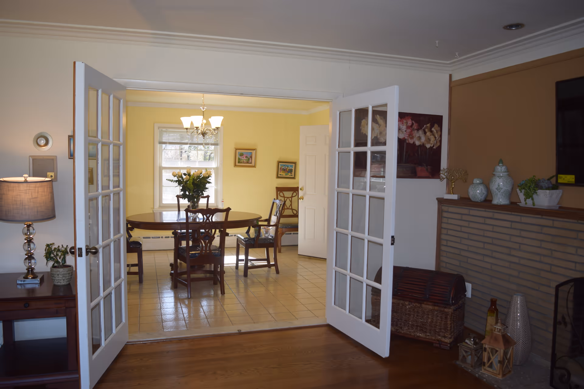 Open French doors reveal a bright dining room with a round table, wooden chairs, and a chandelier.