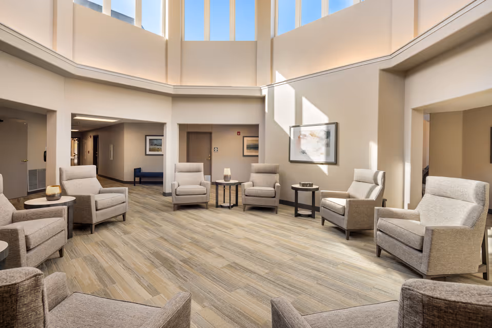A bright and spacious senior living community common area with high ceilings and tall windows letting in natural light. The room features a circle of beige upholstered armchairs arranged around small round tables on a wood-patterned floor. Neutral-colored walls are decorated with framed artwork, and doorways lead to adjacent rooms and hallways.