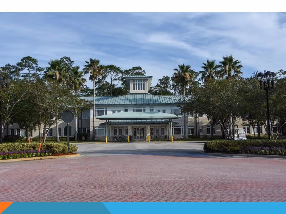 Front exterior view of Westminster St. Augustine facility showing a large building with a green metal roof, multiple windows, and a covered entrance. The building is surrounded by palm trees, other greenery, and a circular driveway with a brick pattern under a partly cloudy sky.