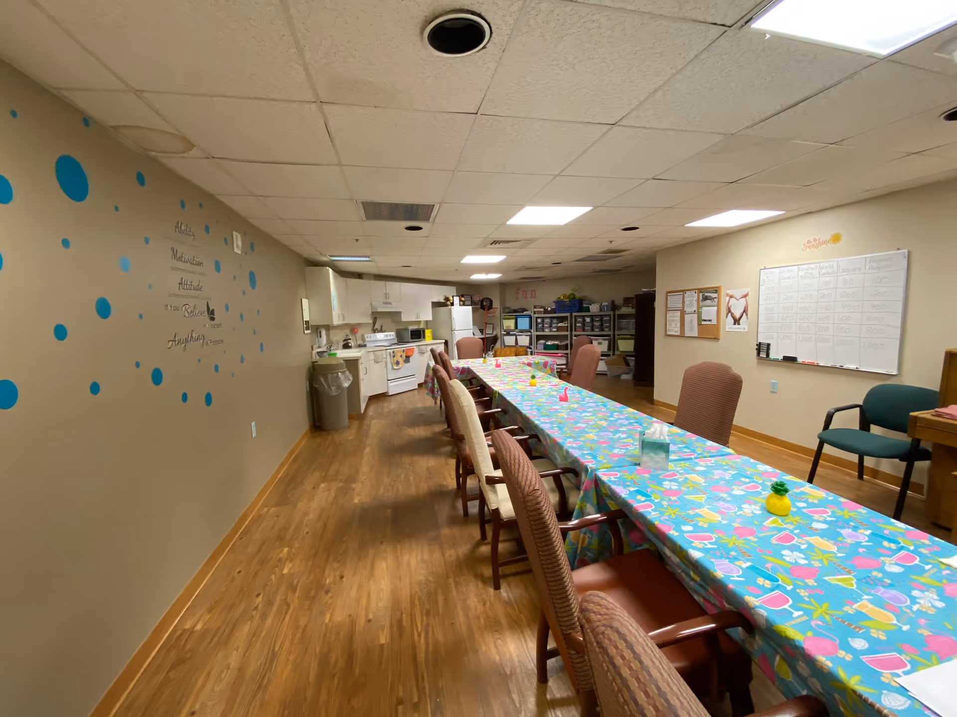 A long room with a wooden floor featuring a table covered with a colorful tablecloth and surrounded by chairs. The back wall has a small kitchen area with white cabinets, a stove, microwave, and refrigerator. The left wall has blue polka dot decorations and an inspirational quote. The right wall has a whiteboard with writing and a bulletin board with papers pinned on it. The ceiling has fluorescent lights and ceiling tiles.
