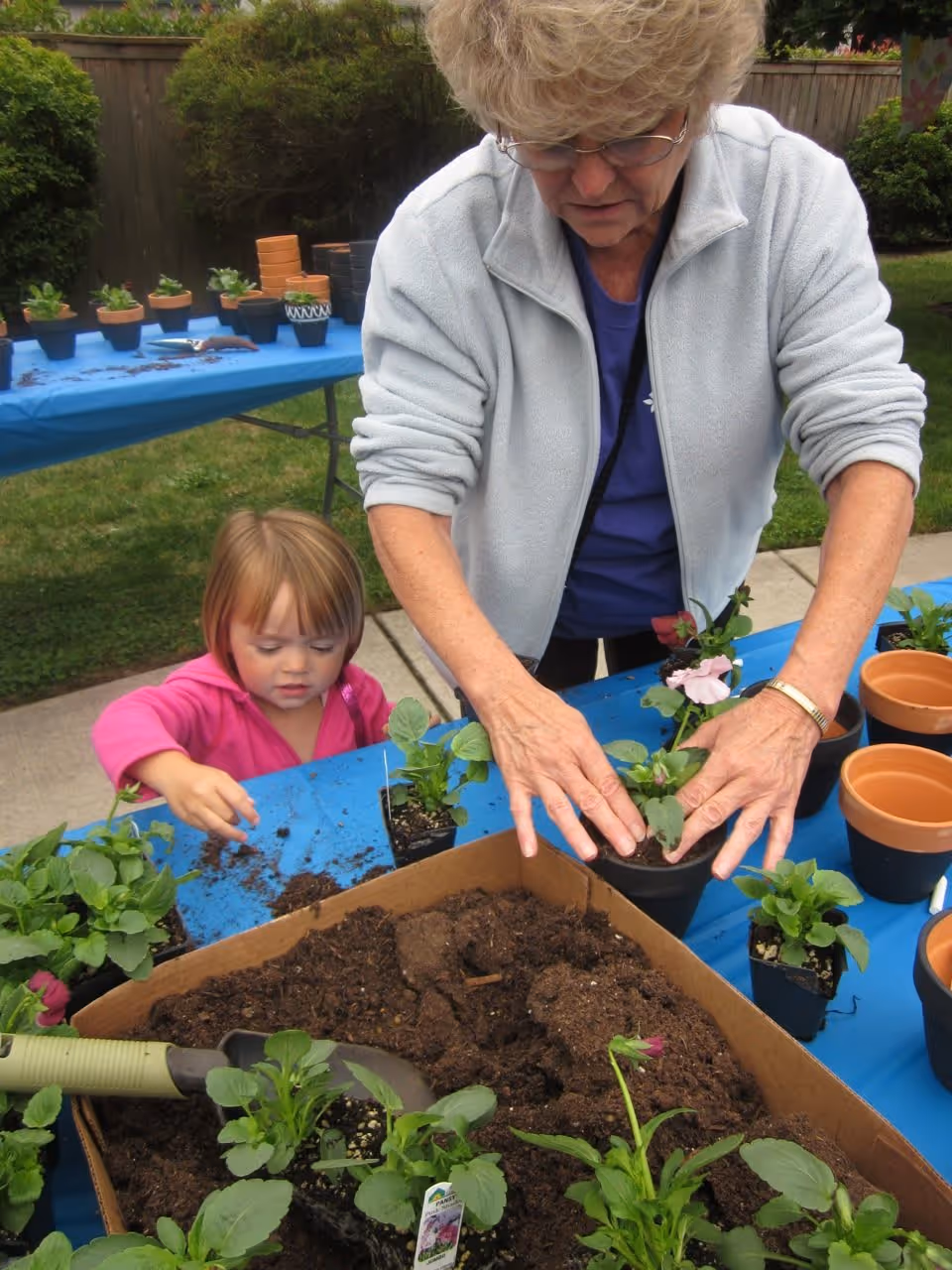 An adult and a young child potting flowers at outdoor tables with soil, seedlings, and clay pots.