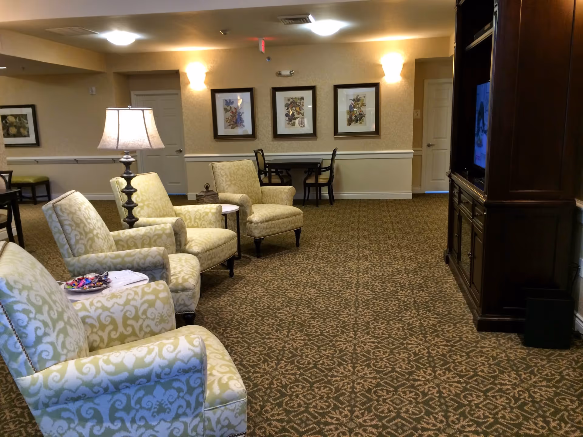 A comfortable senior living common room with patterned armchairs, a floor lamp, framed artwork on the wall, and a TV cabinet.