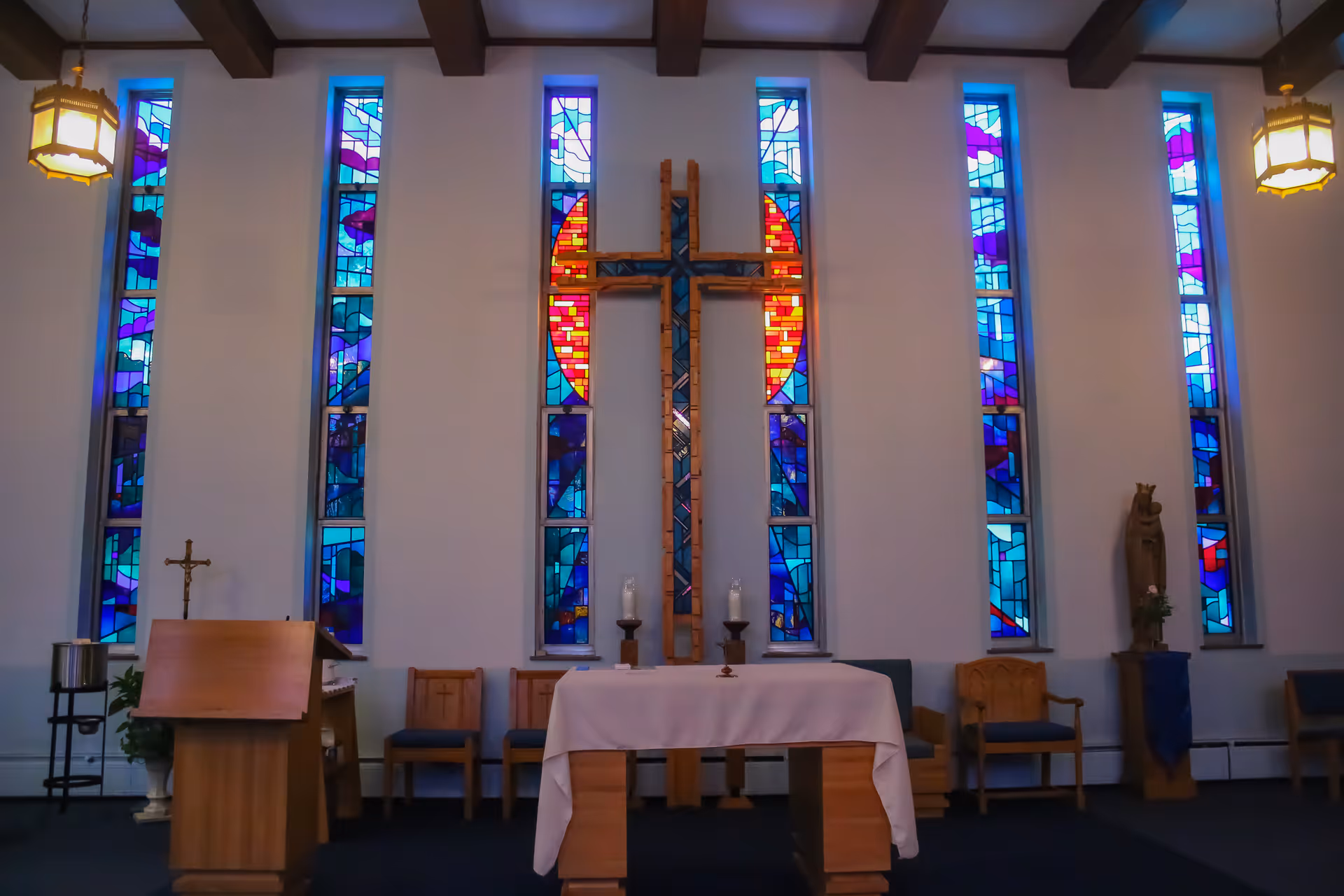 Interior view of a chapel with a large wooden cross mounted on the wall, surrounded by tall stained glass windows featuring blue, purple, and red patterns. Below the cross is an altar covered with a white cloth, flanked by wooden chairs and religious statues. The ceiling has exposed wooden beams and hanging lantern-style lights.