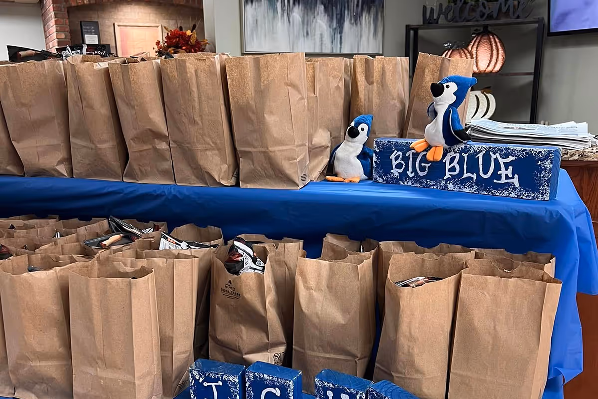 Rows of brown paper gift bags on a blue-draped table inside a facility, with penguin plush toys and a 'BIG BLUE' sign.