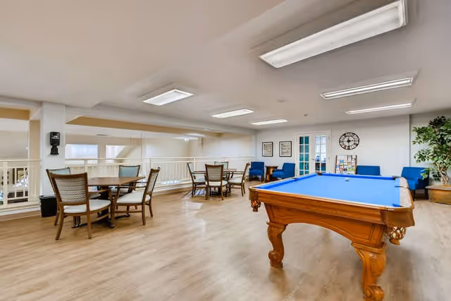 A spacious recreational room with a blue felt pool table in the foreground, several round tables with chairs, blue armchairs along the back wall, a large clock, framed pictures, and a potted plant. The room has wood flooring, white walls, and bright overhead lighting.