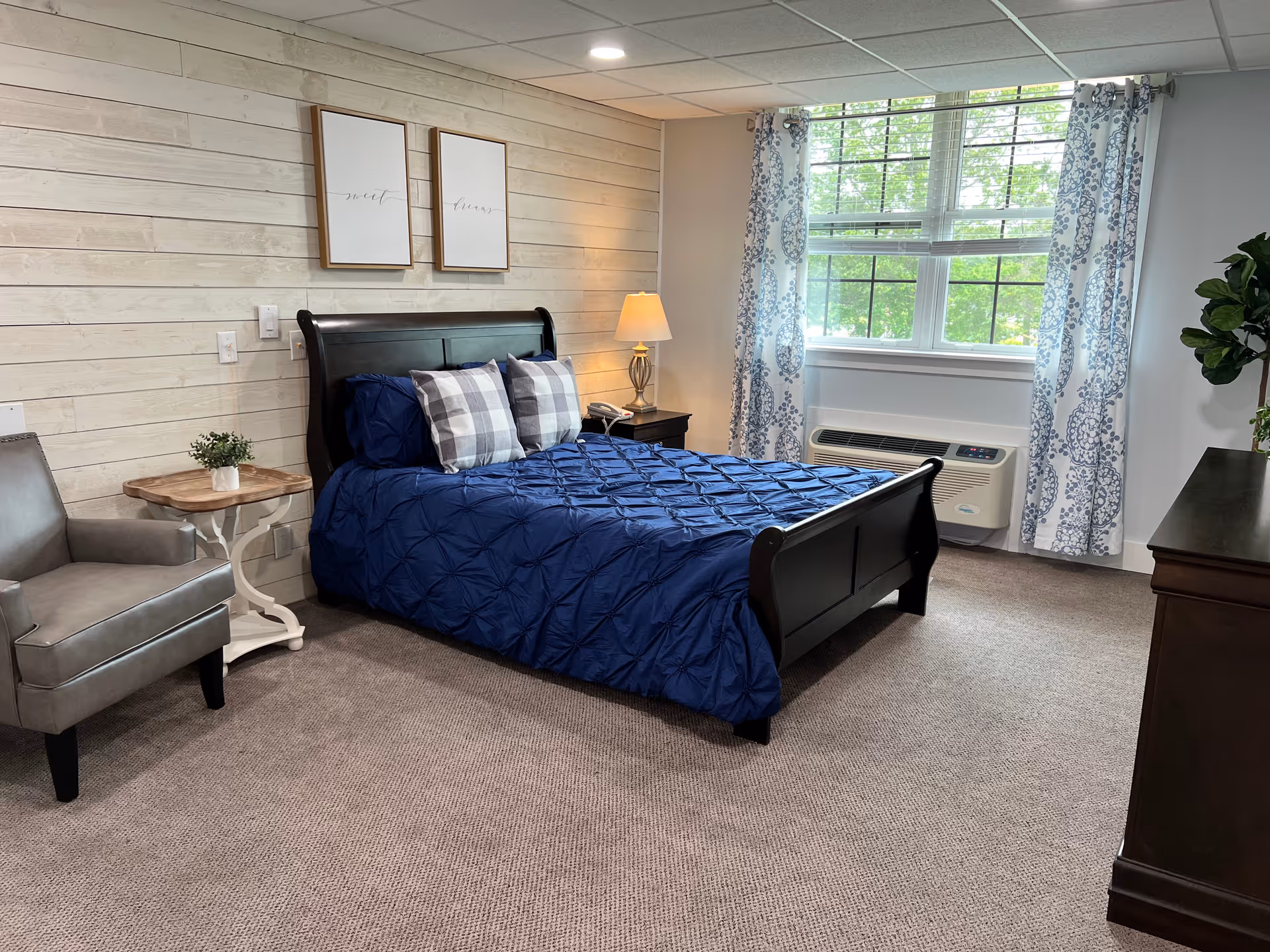 A cozy bedroom with a dark wooden bed frame and a blue comforter. Two plaid pillows and two dark blue pillows are arranged on the bed. There is a gray armchair next to a small wooden side table with a small plant on it. A nightstand with a lamp and a phone is beside the bed. The room has a large window with patterned curtains, a beige carpet, and light-colored wooden panel walls. A dark wooden dresser and a green plant are also visible.