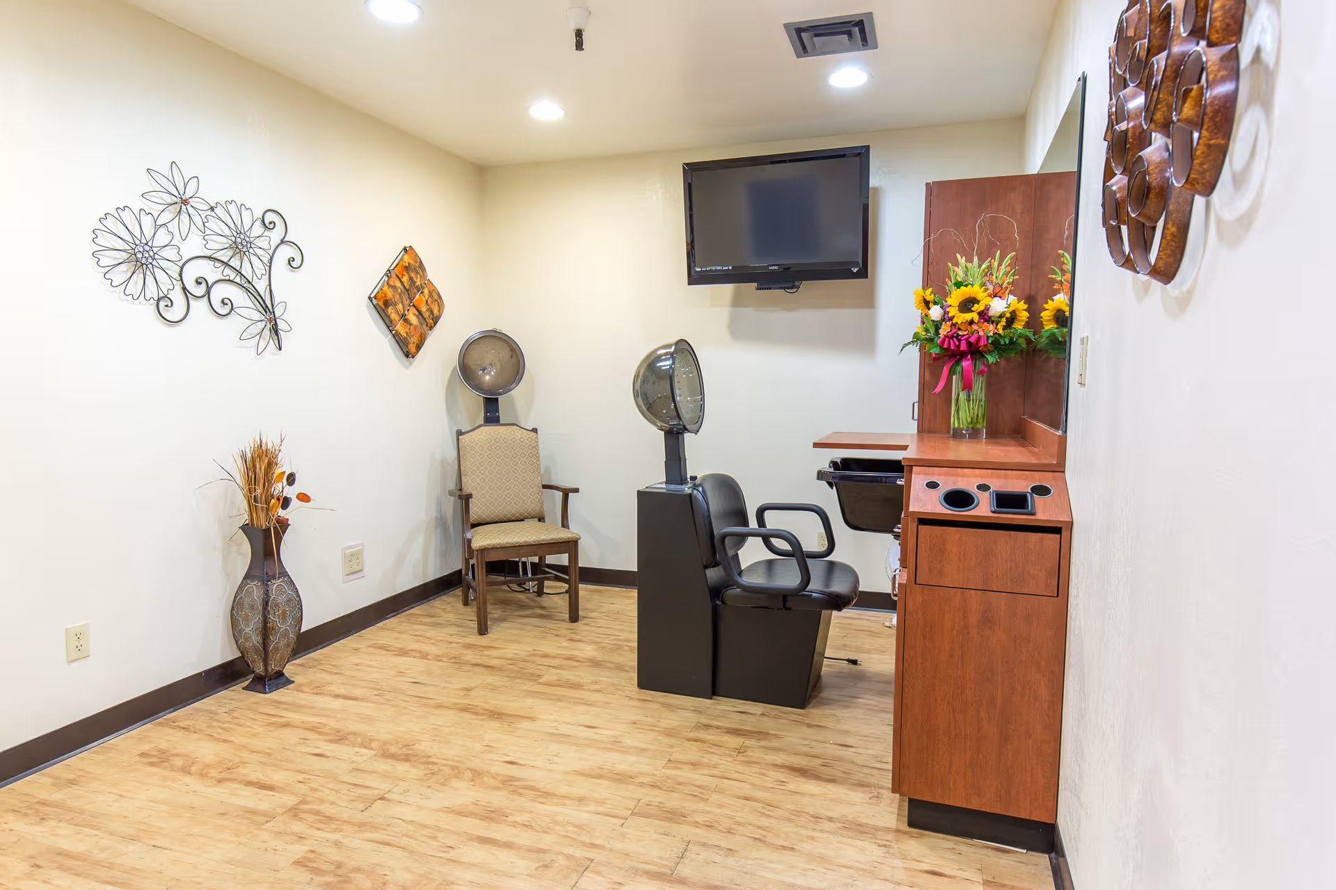 Interior view of a small salon area in a care center with a hair drying chair, a regular chair, a wall-mounted TV, a wooden cabinet with a mirror and a vase of flowers, and decorative wall art.