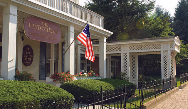 Exterior view of The Easton Home building with a porch, an American flag, manicured bushes, and a white lattice structure extending from the porch.