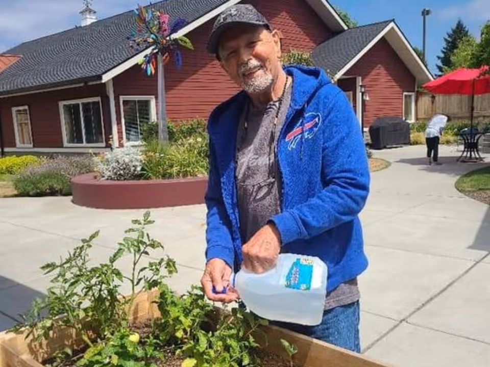 An elderly man wearing a blue jacket and cap is watering plants in a raised garden bed outdoors at a senior living facility. Behind him is a red building with white trim, a colorful wind spinner, and another person tending to the garden near a table with a red umbrella.