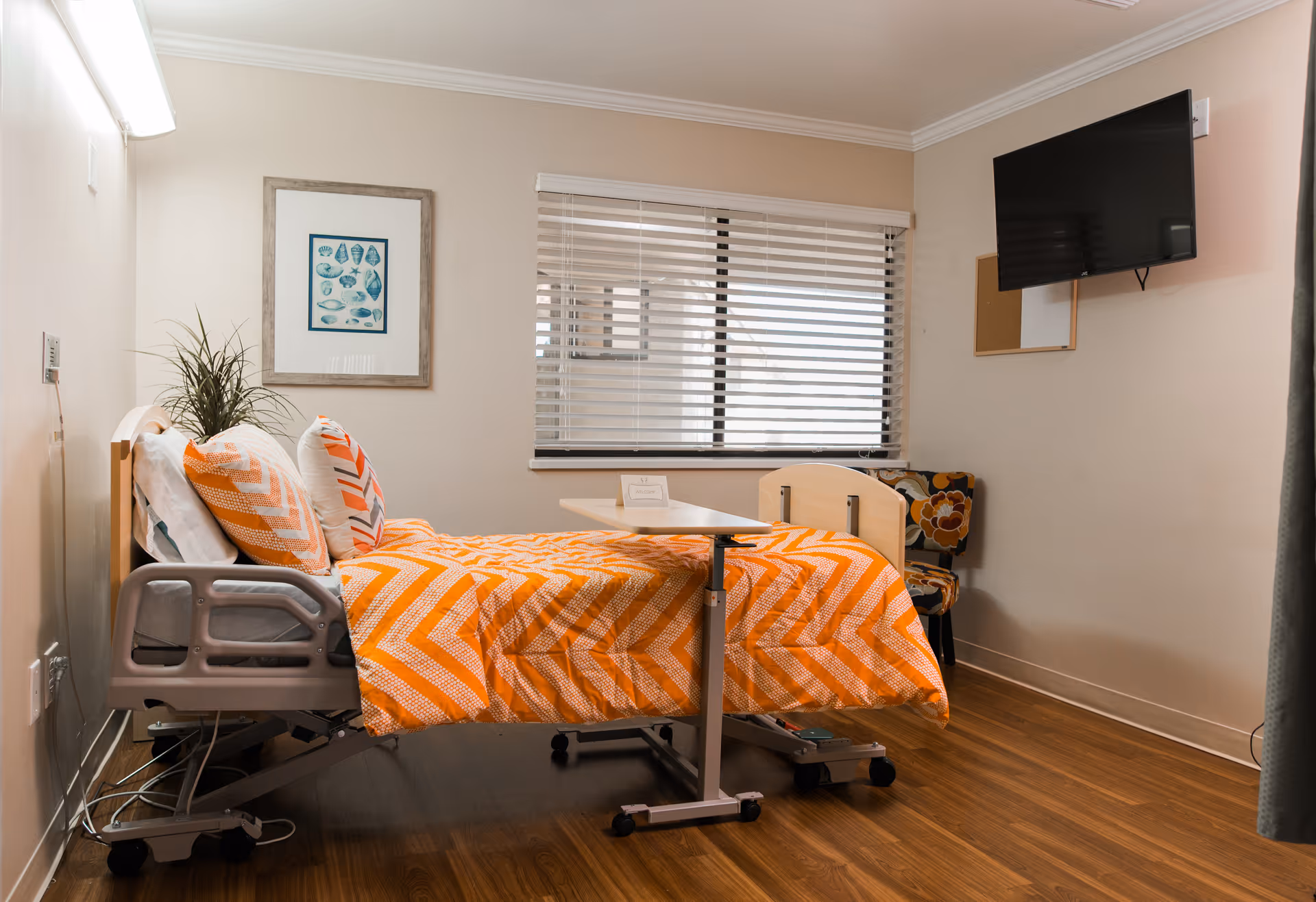 A bright senior-care bedroom with a hospital-style bed dressed in an orange chevron blanket, overbed table, wall-mounted TV, and window blinds.