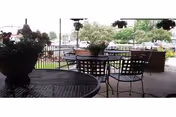 Outdoor patio area with metal tables and chairs, potted plants on the tables and hanging above, overlooking a parking lot and greenery in the background.
