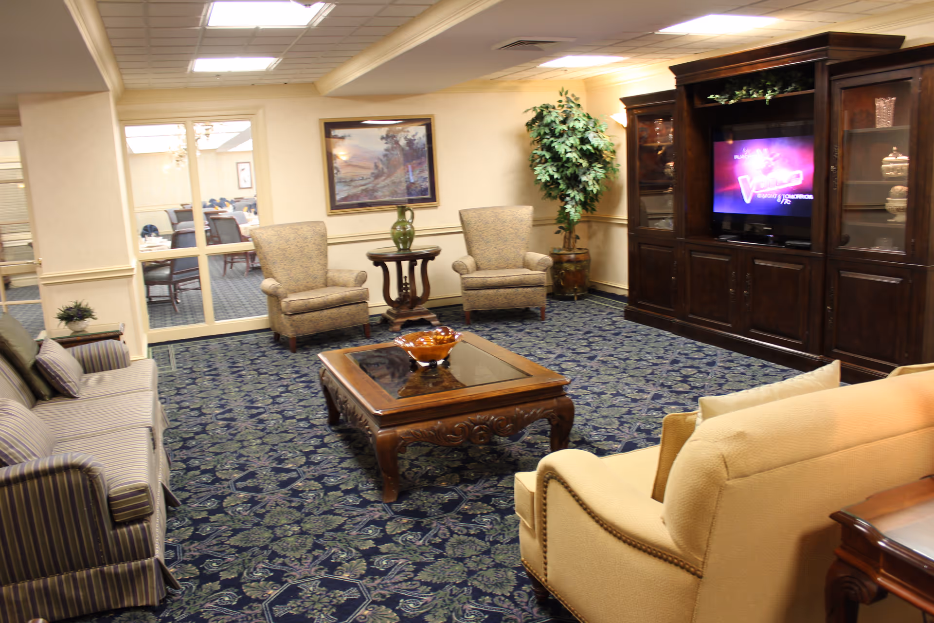 A cozy senior living community lounge area with patterned carpet, two beige armchairs, a striped sofa, a cream-colored sofa, a wooden coffee table with a decorative bowl, a large wooden entertainment center with a TV, a potted plant, and a framed landscape painting on the wall.