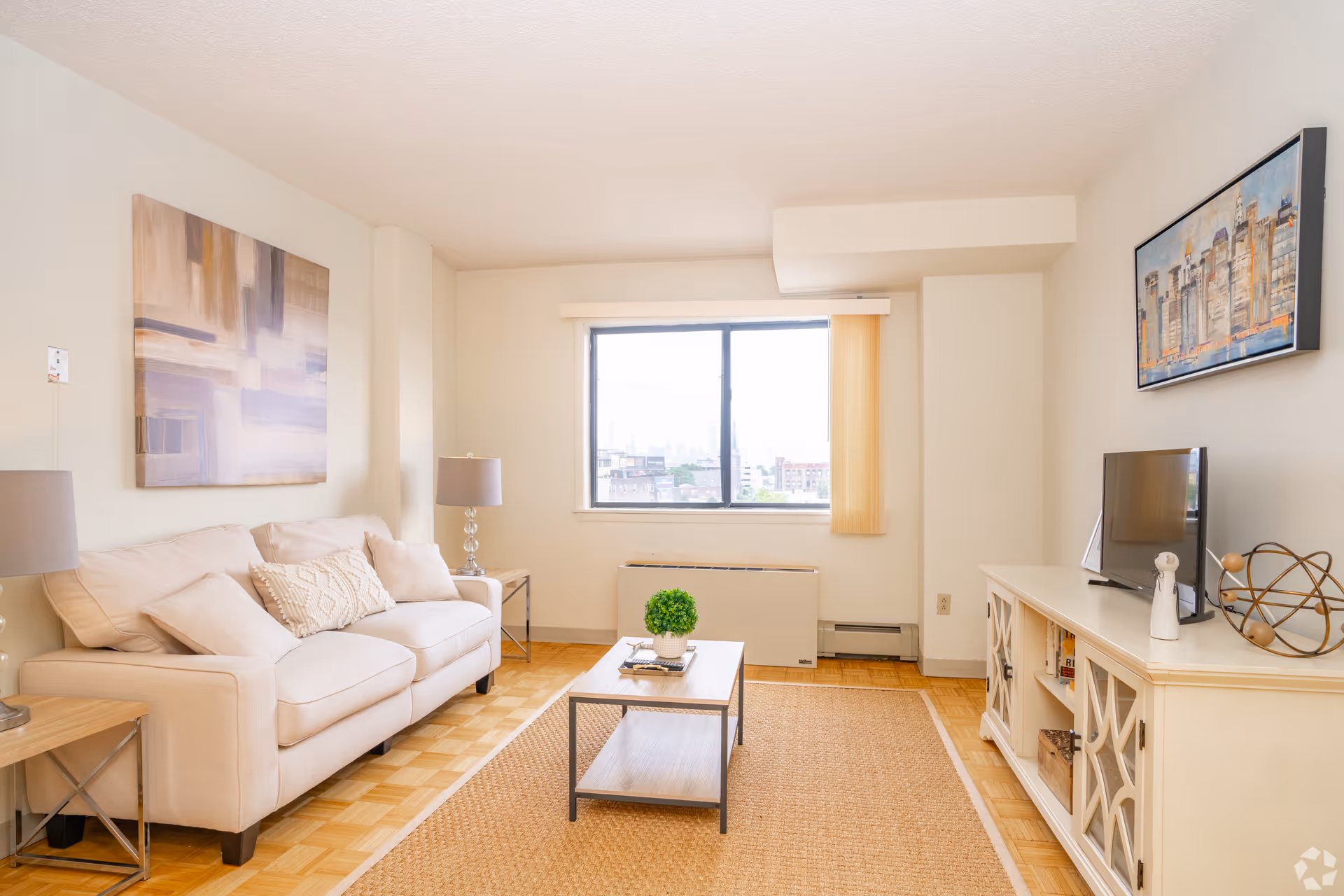A bright living room with a beige sofa adorned with cushions, two side tables each with a lamp, a rectangular coffee table with a small green plant, a large window with vertical blinds, a TV on a white cabinet, and abstract artwork on the walls.